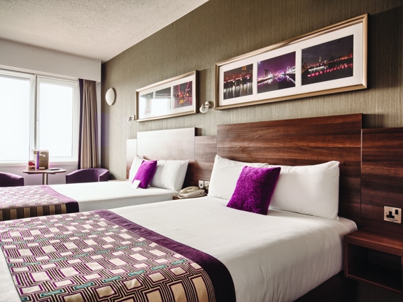 A view of a Leornardo Hotel twin room, 2 single beds dominate the image, the dark wood-effect headboard takes up the length of the visible, right-hand wall. The beds are made with crisp white sheets and a geometric patterned throw in shades of purple and teal. The far corner has a curtained window, in front of which a small, round bistro table and two mauve fabric chairs are positioned. A telephone, multiple plug outlets, framed prints and light fixtures are also all visible.