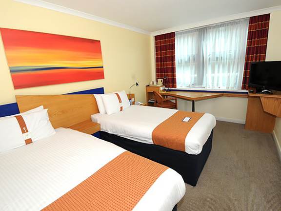 An interior image of a Holiday Inn Express twin room shows a room with a neutral, textured carpet and cream walls. 2 beds are made up with crisp white sheets and a narrow orange throw. An angular wooden desk and TV stand takes up the furthest wall. There is a wide window with white net curtain, heavier tartan curtains in shades of red and pulled back. An abstract print in shade of red and orange is above the beds. 