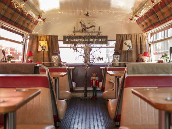 A photo of the Red Bus Bistro's upper deck, decorated for Christmas. Vintage leatherette bus seats, in red and tan, have been modified to face one another over small wooden topped tables. The photo is taken from half way down the bus, looking towards the wide,  top, front window. A silver-birch branch at the end of the central aisle is decorated with baubles, and fairy lights line the ceiling. There are small gingham curtains on the front window and tartan upholstered padded panels on the ceiling.