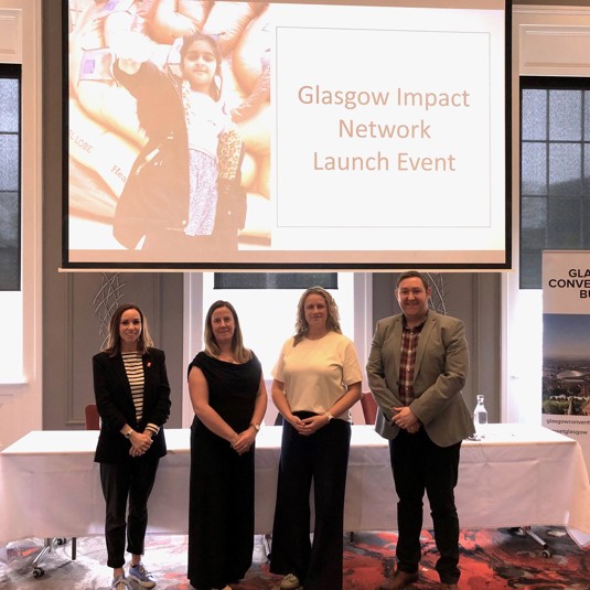 Helen Davidson, Dr Lorraine Work, Dr Deborah Mitchell and Campbell Arnott standing together smiling in front of a long table with a presentation screen above them which reads 'Glasgow Impact Network Launch Event'