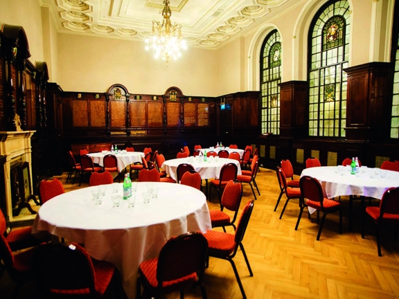 A photo of the Trades Hall room, The Saloon, with a varnished, parquet floor and high ceilings. The walls have dark wooden panelling to half the room's height, the left and far walls have golden writing and coats of arms further decorating the panels. 3, huge, arched stained glass windows decorate the right wall, a marble fireplace adorns the wall opposite. Round tables with white table cloths & chairs with red upholstery are arranged in groups. A lit chandelier hangs from a decoratively plastered ceiling.