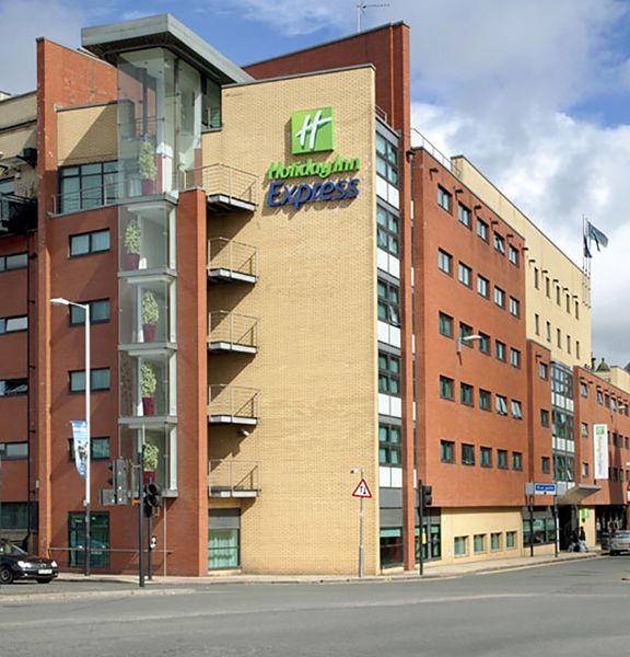 An exterior image of the Holiday Inn Express Riverside shows a modern yellow and red brick multi-storey building, from the viewpoint it is possible to see the building is on on the corner of 2 roads. A large sign on the end of the building reads "Holiday Inn Express" in the brand's green and blue. An entrance can be seen down the right-hand road, with a white sign marking it. The building is seen against a blue cloudy sky.