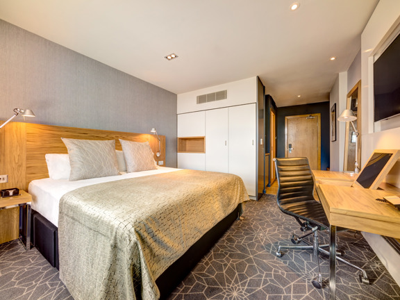 A standard double room at Apex Glasgow. An interior shot of a hotel room decorated with grey, patterned carpet and wallpaper. The bed is large and tidy and is decorated with a grey blanket and cushions. There is a television, desk, chair and mirror all visible.