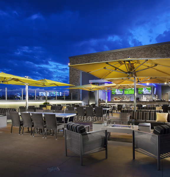 An image of the TopGolf Terrace Bar during the evening. A large paved balcony area is filled with cushioned, metal sofas, woven high-backed chairs, polished stone tables and large, square, yellow parasols. The light-brick building attached, has 2 huge cut away walls to the interior. A stone-effect bar, large TV screens and further seating can all be seen within. 