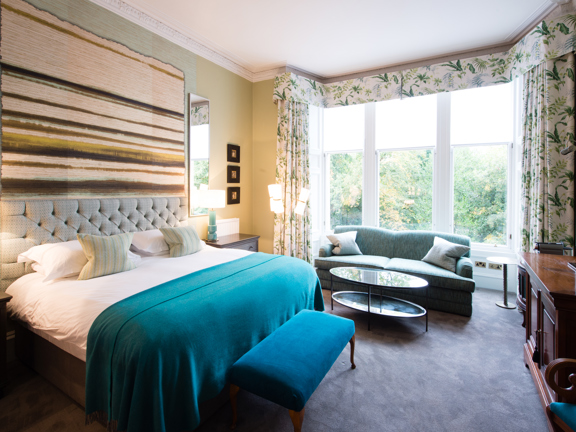 An interior view of one of Hotel du Vin's bedrooms, the room has soft grey carpet and very pale green walls. A large bay window is adorned with white curtains with a green leaf print. A pale turquoise sofa and a low, mirrored coffee table sit in it. A large double bed sits against the left wall it has crisp white sheets and a teal throw, the headboard is padded and beige. Above the bed is a large textile weaving in creams and browns. Against the right wall is a dark wood dresser with a large TV on top.