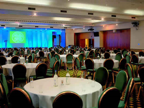 An internal view from Celtic Football Club shows a large hall with a dark, patterned carpet. The room is filled with round tables covered in white table cloths, surrounded by plush, green chairs. A stage is visible on the far wall, it is brightly lit with green and turquoise lighting and a lectern and speakers are visible.
