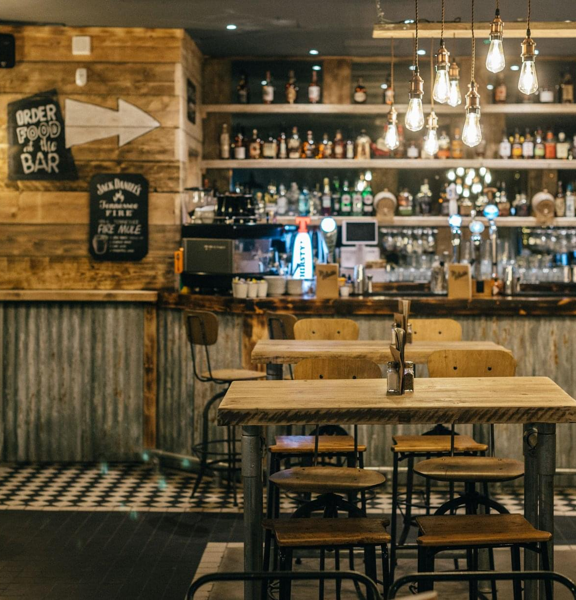 An image from inside the Van Winkle restaurant and bar, shows a room with rustic, reclaimed wood & corrugated metal panelling on the walls. The floor is tiles in a mix of black and white. High wood and metal stools sit at high scaffolding-style tables and counters. Bare, filament lightbulbs with brass fittings hang in groupings. A well stocked bar and shelves takes up much of the back wall, and small wall mounted blackboard signs list offers and direction.