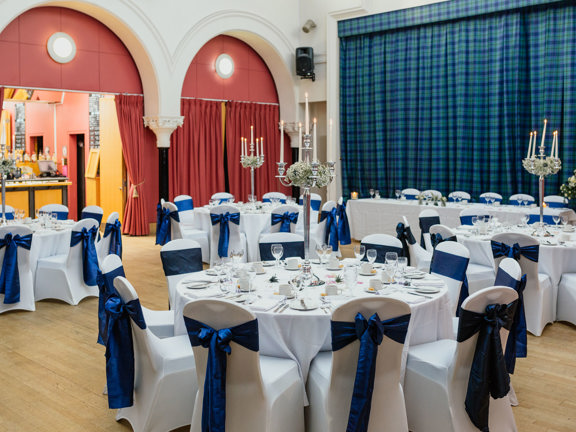 An image of a private event set-up at The National Piping Centre. A hall with wooden floors and a stage curtained off with a heavy green and blue tartan carpet on the right hand wall. Round tables are covered with white table cloths and set with silverware, glasses and tall, silver candelabras. Chairs are covered with white covers and decorated with wide, royal blue ribbons. In the far wall 2 arched openings are visible; red curtains pulled back on one of them reveals a brightly lit pine bar.
