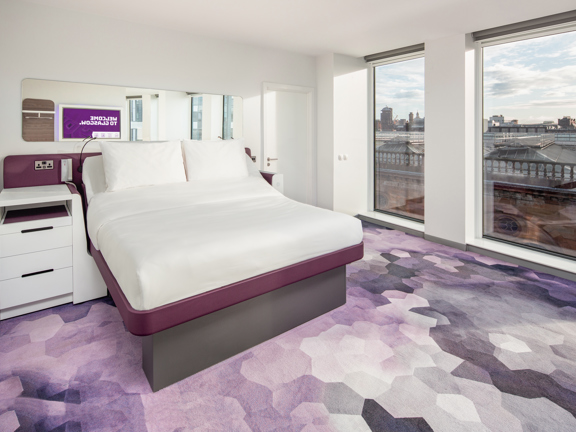 Yotel's modern, minimal "cabins" bedrooms, overlooking the Victorian glass and steel roofline of Central Station and red sandstone balusters, through the large plate glass windows. The room is carpeted in an purple ombre honeycomb. The bed is made with crisp white sheets and a white bedside table sits on the left hand side of the bed. On the right of the bed is a closed doorway and in the long mirror over the bed a television screen can be seen on the opposite wall.