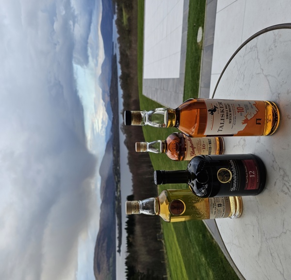 Four bottles of whisky sitting on a table with a view of Scottish mountains and loch behind