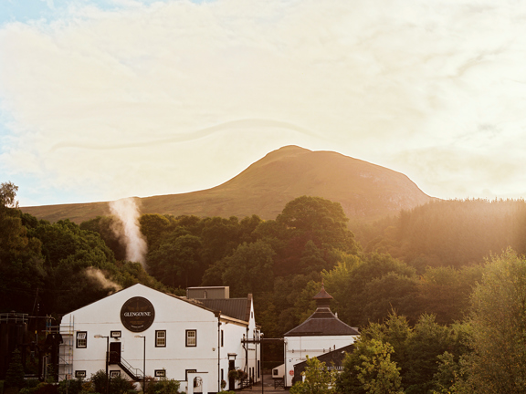 An exterior view of the Glengoyne Distillery shows a 3 storey building with white rendering and a gabled roof, dwarfed by the trees and hills behind it. There is a neighbouring smaller building obscured by trees. The large building has a large black circle on it with white lettering inside it reading "Glengoyne." The sky is bright giving a golden glow to the large trees, the striking outline of a hill and the sky. It also highlights the steam or smoke rising from the main building. 