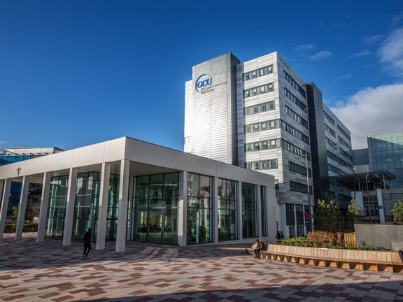 An external shot of Glasgow Caledonian University shows a paved path of different coloured stone leading to a modern, square silver building about 8-storeys high. At the top of the front facade large, blue lettering reading "GCU" can be seen; smaller white lettering is visible but hard to read. There is a single storey, glass entrance atrium in the foreground. To the right of it is landscaped flower beds and a long curving wooden bench lining the paved area.
