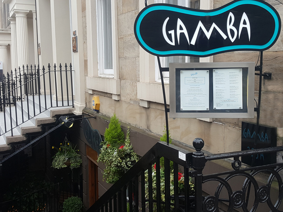 An exterior view of Gamba shows a glossy, black wrought iron stairway leading to the basement floor of a Victorian sandstone building. A rounded black and turquoise sign is visible above a framed menu. White, angular lettering reads "Gamba". Hanging baskets filled with ivy, geraniums and miniature yews decorate the outside of the restaurant. The windows of the upper floor and stairs, railings and pillars of the neighbouring building's doorway are also visible.