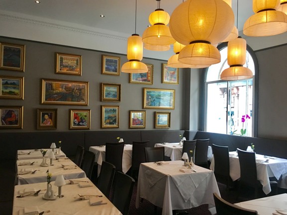 An image of the Amber Regent private dining space. A modern dining room with grey upholstered seating around the edge of the room, small tables with while table cloths and high-backed grey chairs. The room is decorated with small paintings in gilt frames and tissue paper lampshades in different shapes and sizes hanging from the ceiling In the corner, daylight streams through an arched window, a pink orchid sits on the window sill.