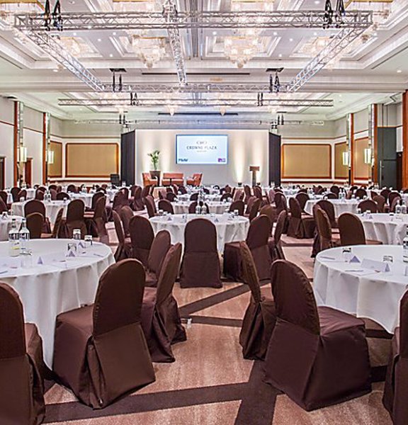 An interior view of the Crowne Plaza ballroom shows a large room decorated in neutral tones of brown and beige. The floor is filled with large round tables, covered with white table cloths and chairs around them with brown fabric covers. At the far end of the room a stage with 3 seats and a podium is evident, behind which a projection screen is pulled down. There are chandeliers in the ceiling as well as a technical light rig. 