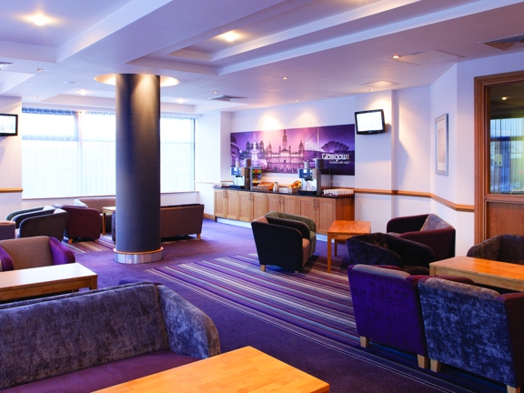 An interior view from the Leornardo hotel shows the meeting space, breakout room. Small groupings of low, purple and grey armchairs and sofas surround wooden coffee tables. The floor is a striped carpet in the same tones and the walls and ceiling are a bright white. Windows with vertical blinds line the far, left wall. A dark, cylindrical pillar obscures the view of a catering bar, where 2 coffee machines and a fruit bowl are just visible. A small, wall mounted TV can be seen on either wall.