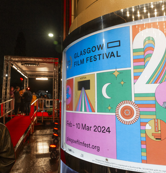 A person with their back turned stands next to the red carpet and a colourful poster for Glasgow Film Festival 2024.