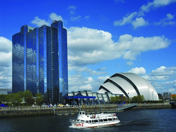 An external view of the Crowne Plaza hotel evidencing the hotels proximity to the Scottish Event Campus and the River Clyde. In the foreground a white boat passes the hotel - a glass, high-rise building. Next to the Crowne Plaza is the famous SEC "Armadillo" building, a silver segmented structure, shines in the sun. Other buildings and trees are also present.