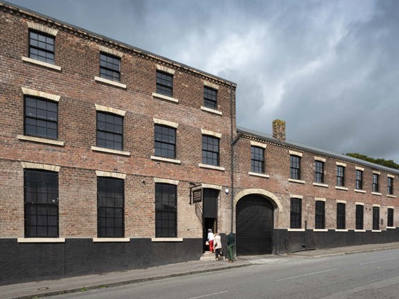 A view of The Engine Works building's exterior, lining the far side of a quiet street with narrow tarmac pavements. 3 people are climbing the small stone steps into the main entrance. To their right is a large arched entrance way with a black shutter. The building is mainly red brick with black and white brick accents. The roof is stepped with the building's left half boasting 3 storeys and the right half only 2. Large, paned, black framed windows line both halves. The roofline is framed against a grey sky.