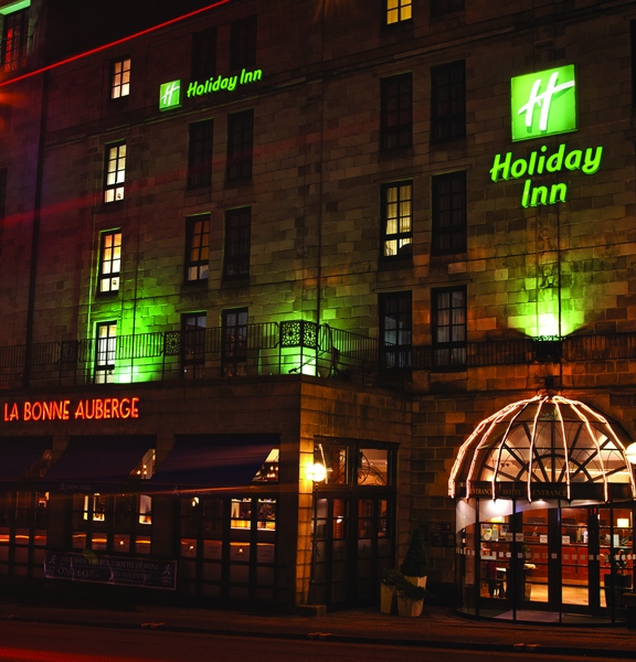 An exterior view of Holiday Inn Theatreland by night shows a 6 storeyed stone building. The building is uplit on the first floor with green lights and 2 lit signs on the facade read "Holiday Inn" and bear the companies logo in green. The main entrance is a glass, sliding door, with an arched window above. On the ground floor a space with large, windows and awnings juts out from the rest of the building; it has red neon signage reading, "La Bonne Auberge." To the left streetlights and buildings are visible.