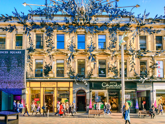 An image of the exterior of Princes Square from Sauchiehall Street. A 3-storey blonde sandstone building on a pedestrianised street. The building's entire façade is symmetrically decorated by intricate, botanical, modern ironwork. Different businesses take up the ground floor, glass plated shop spaces. On the left, a large LED screen displays 'Princes Square' in a serif font above a black and white geometric spray of lights. It is a sunny day and pedestrians, lamposts and benches can be seen.