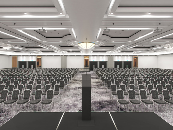 An interior view from the front a large event space at Hilton Glasgow shows a large, bright, carpeted room. The floor is filled with dozens of dark grey chairs arranged into roughly 20 rows. In the foreground a small, temporary stage and a slim metalic lectern are centrally aligned with a large, domed glass light feature in the centre of the room. Projectors can be seen mounted to the finely-panelled ceiling intermittently. 6 black double doors and large, gold-framed mirrors dot the far wall.
