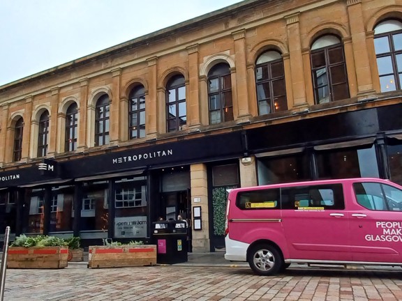 An exterior of Metropolitan, cocktail bar and restaurant, shows a long 2 storeyed sandstone building. The first floor is lined with dark-framed arched windows the ground floor has large floor to ceiling windows and a black sign with white lettering reading "Metropolitan". The road surface looks cobbled but the pavement is paving slabs. A pink, 8 seater taxi, public bin, bollards and planters can all be seen on the street. On the right, a large arched entrance way can be seen.