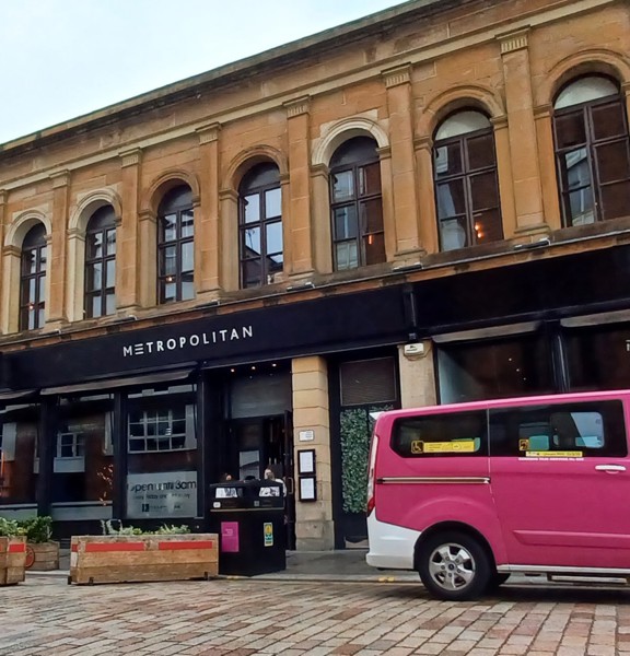 An exterior of Metropolitan, cocktail bar and restaurant, shows a long 2 storeyed sandstone building. The first floor is lined with dark-framed arched windows the ground floor has large floor to ceiling windows and a black sign with white lettering reading "Metropolitan". The road surface looks cobbled but the pavement is paving slabs. A pink, 8 seater taxi, public bin, bollards and planters can all be seen on the street. On the right, a large arched entrance way can be seen.