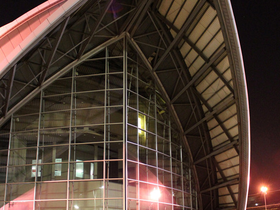 A large, black, shiny people carrier parked outside one of the pointed ends of the SEC armadillo building. The image is taken at night lit by bright lampposts and the car headlights. The building is modern and glass.