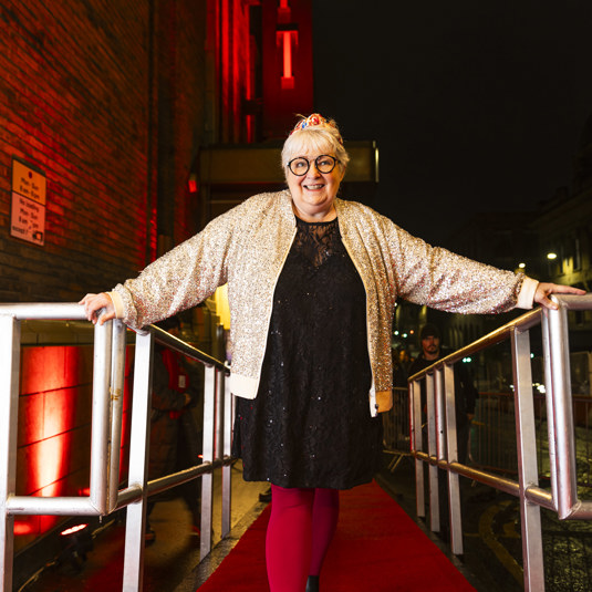 Janey Godley poses in a black dress and glitter jacket on the red carpet, outside the Glasgow Film Theatre.
