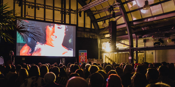 A crowd face a big screen. The screen displays a person with their mouth open.