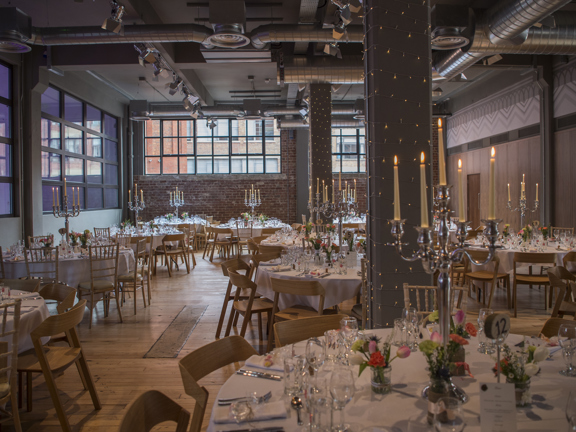 A West Brewery space arranged and decorated for a private dinner. The space has a post-industrial aesthetic, with bare brick walls, girders, and silver extraction tubes on the ceiling. The room is decorated with fairy lights and the several round tables with crisp white table cloths, tall silver candelabras and small posies of flowers. The chairs are wooden and a mix of shapes. The floor is a polished, reclaimed wood finish. Large, landscape windows with dark framed cover the top half of the far wall.