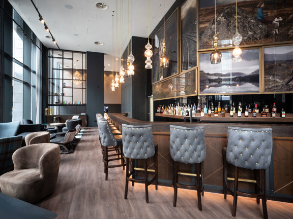 An interior shot of Motel One's bar shows a long, narrow room between a floor to ceiling, external window on the left and a long wooden bar on the right. The floor is an ashen wood and the walls are grey and decorated with framed landscape photos. There is a mix of high seats at the bar and comfortable, low chairs and tables positioned along the window. Modern, glass lights hang over the bar and the shelves behind are well stocked with bottles.