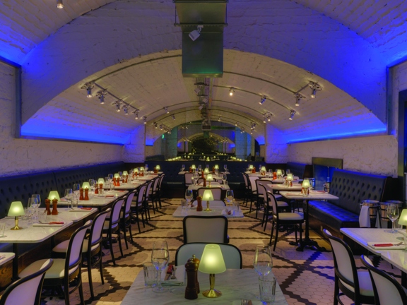 An interior of the Alston Bar & Beef's underground dining room. The walls and curving ceiling of the room are all white-painted brick, blue strip lights up-light the walls and small, white spotlights mounted to the ceiling provide lights. The edges of the room are lined with leather upholstered benches, white marble tables laid for dinner and dark, wooden chairs with cream upholstery furnish the space. The floor is tiled with small black and white tiles laid in geometric shapes.
