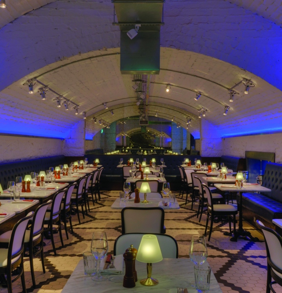 An interior of the Alston Bar & Beef's underground dining room. The walls and curving ceiling of the room are all white-painted brick, blue strip lights up-light the walls and small, white spotlights mounted to the ceiling provide lights. The edges of the room are lined with leather upholstered benches, white marble tables laid for dinner and dark, wooden chairs with cream upholstery furnish the space. The floor is tiled with small black and white tiles laid in geometric shapes.