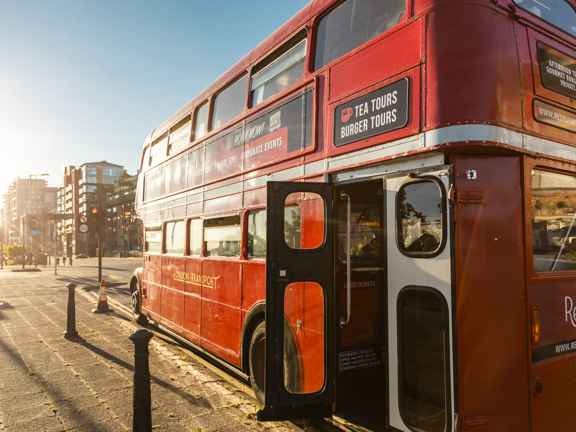 A red, vintage double-decker bus parked on a sunny day. Panels on the side and back advertise the bistro. The bus is parked near a junction with traffic lights, a green space is visible over the road. The pavement is level and has a curb.  The doors at the back of the bus are open, revealing a small step up. The sky is blue and the sun is shining. 