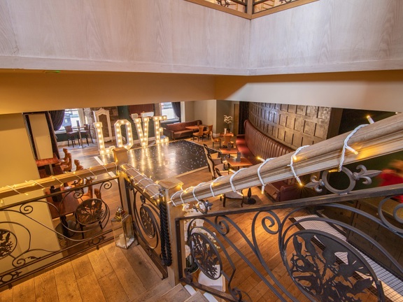 An interior view from Citation from the top of a wooden staircase. The staircase leads into a room with wooden floors and panelled walls. A long, plush bench runs the length of a curved wall, other tables and chairs dot the room. The staircase banister is decorated with fairy-lights and in the centre of the space a tiled dancefloor and a large light up sign that reads "love" dominate the space in front of two large windows.