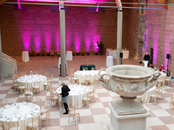 An internal view of the main foyer of The Burrell Collection set up for a dinner event. A large space with a checkerboard stone floor. Round tables with white table cloths surround the Warwick Vase, a large, marble urn. The far wall is red sandstone and up-lit with pink lights. A lectern, speakers, plants and staff-members are also visible.