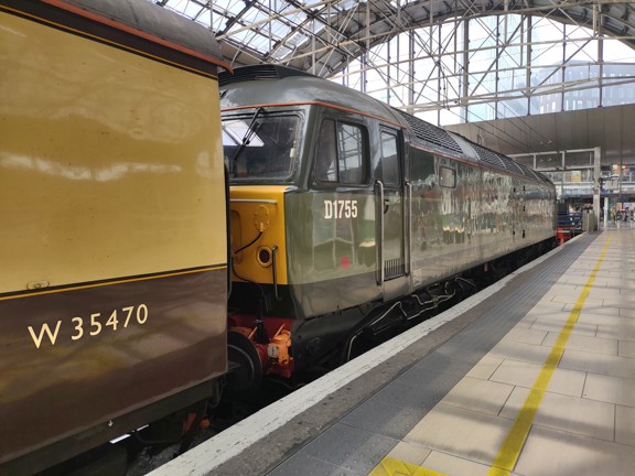An old diesel train in its platform at Queen Street station. The train is blocky and painted olive green,  the end of an old train carriage is visible in front, painted brown and cream.  The platform is paved and level, with a row of tactile tiles, a white edge and painted yellow guidelines. The arching glass roof of the station and concourse area are visible above and beyond.