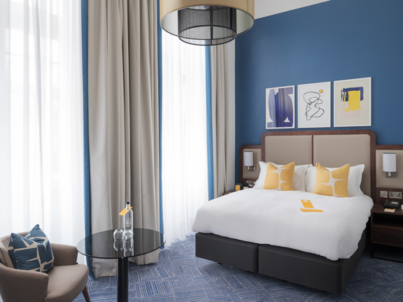 A voco Grand Central double room shows a stylish, modern hotel rooms with blue walls and cream carpet with a blue crosshatch pattern. The bed headboard and bed side tables are made from the same piece of dark wood, with padded panels in a taupe fabric. The bed is made with crisp white sheets and has yellow, accent cushions. A small modern cocktail chair and glass coffee table sit at the end of the bed. 2 blue and yellow abstract prints are framed above the bed. The left wall is dominated by 2 huge windows.