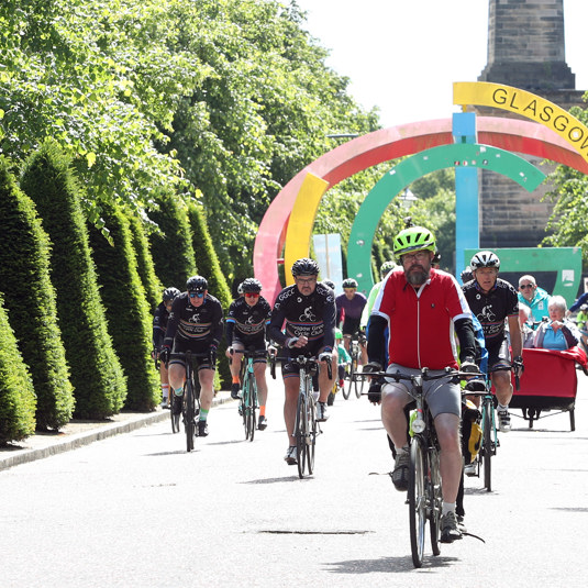 A group of cyclists cycle down a path lined by trees, with a Glasgow 2014 monument behind them.