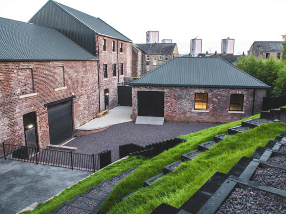 A view of The Engine Works private garden taken from the top of a terraced grassy slope. A set of dark, gravel steps zig-zag down the slope to a triangular yard partly paved but predominantly gravelled. There are 2 brick building with metal rooves off of the yard, a smaller one on the right has a ramp to its door, the lights are on lighting up the dark framed windows. A much larger building on the left, has several entrances visible, each with ramped access to them. Trees and tower blocks can be seen beyond