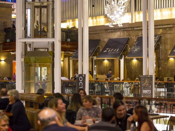 An image of diners in the tiered interior of Princes Square. Black metal railings, a polished glass elevator, white "leafy" light features and restaurant awnings decorate the vast, open space behind them.