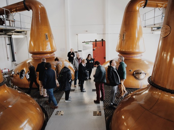 Group of people standing in a whisky still house with giant four copper stills for making whisky