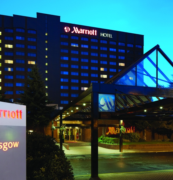 An exterior view of the Glasgow Marriott hotel at night shows a multi-storey square building. Leading to the front door is a long glass covered walkway. In the foreground there is a lit sign reading "Marriott Glasgow" and at the top of the building lit letters read, "Marriott Hotel". Shrubs and landscaping are evident on either side of the walkway.