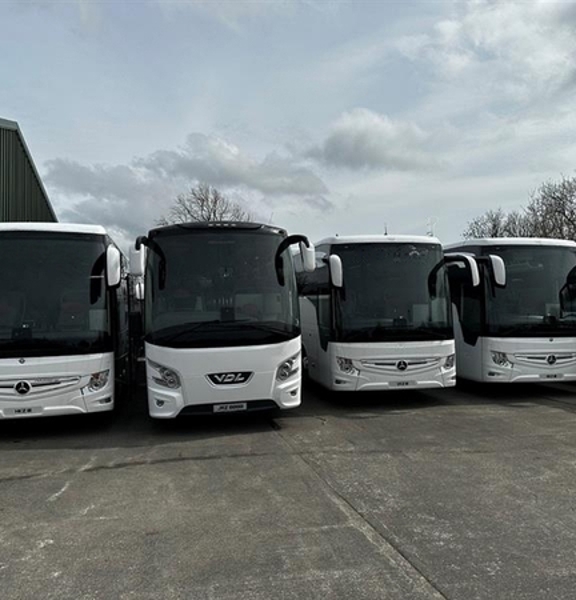 The image shows the fronts of 5 large white coaches parked side by side. On the left of them, the edge of a large grey corrugated building, which could be interpreted as a garage is visible, just on the edge of the frame. Bare trees can be seen beyond the coaches against a pale, cloudy sky.