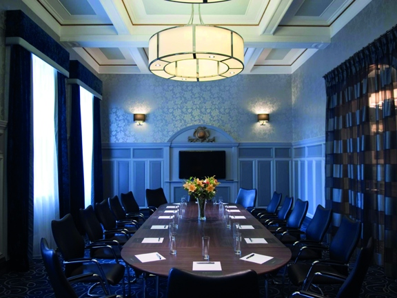 A dark board room meeting space at voco Grand Central. A large oval, wooden table and the surrounding leather and chrome chairs take up a lot of the room. A vase of lilies and a modern, glass ceiling light decorate the space. The room is carpeted with a swirling pattern, the walls panelling is painted pale grey. 2 windows with net curtains across them and heavy dark blue curtains pulled back sit by side on the left hand wall. The right wall has a geometric patterned curtain pulled across an arched doorway.