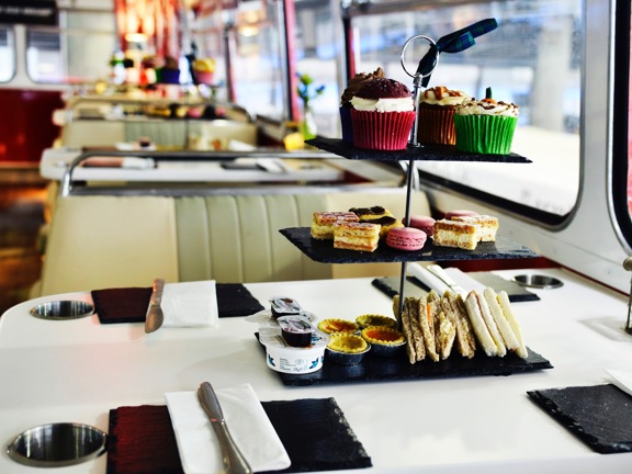 An close up image of an afternoon tea on the Red Bus bistro. A black slate cake stand with 3 tiers is adorned with small cakes in colourful paper cases, triangular, filled sandwiches and macarons. 4 places, with slate plates, knives and paper napkin are set around it. Chrome cup holders are set into the top of the while table top. The vintage leatherette bus seat with chrome edges and another decorated table can be seen to the left of the long right-hand windowed wall.
