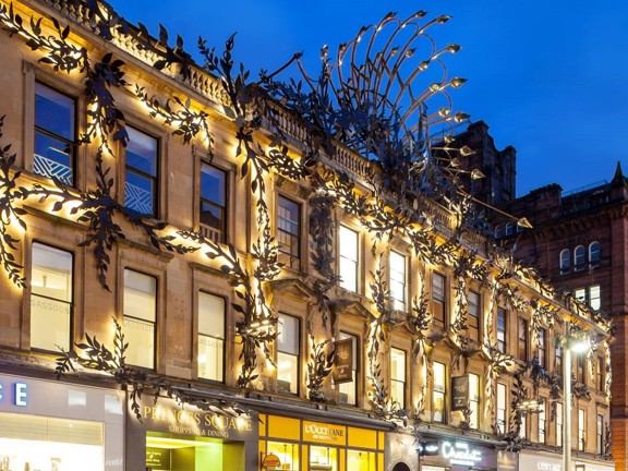 A detail of the intricate, modern iron-work in botanical inspired shapes. It is the evening and the black leaf and feather motifs are lit with golden light. The paved floor of the pedestrianised street is wet with rain; lights from the ground floor shops and decor reflect off the wet street.