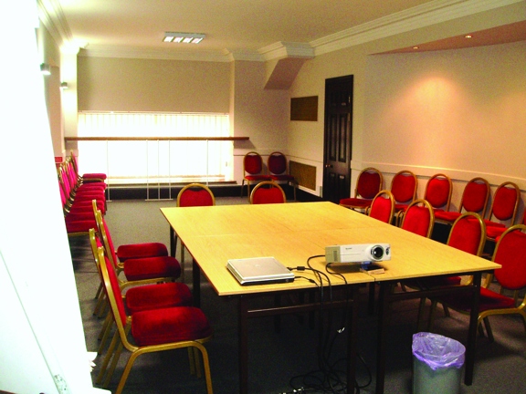The interior of a small meeting room at the Glasgow Trades Hall. The room is carpeted and has cream walls with white plaster beading around the ceiling. A large, low window fills much of the far wall and a dark wooden door enters from the right. The room is equipped with many red upholstered chairs, some surround a set of 4 wood-effect tables arranged into a square. A projector, a bin and the edge of a flip chart are all also visible.